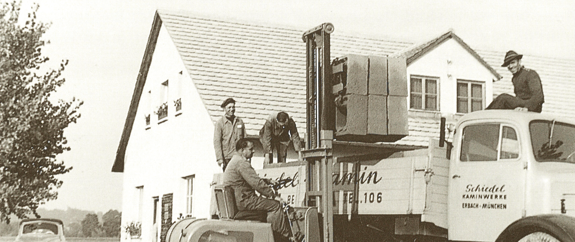 A historic black-and-white photograph showing workers loading heavy concrete chimney blocks onto a vintage Schiedel delivery truck using an early industrial forklift in front of a company facility.