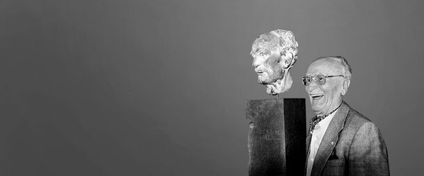 A historic black-and-white photograph of a smiling Friedrich Schiedel standing next to a bronze portrait bust mounted on a dark pedestal.